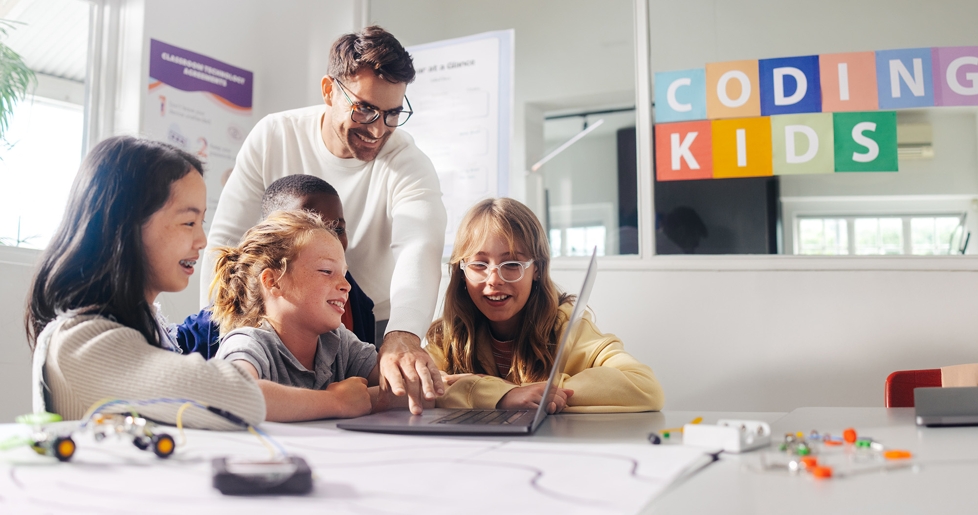Male teacher guides elementary school students in a robot programming class. Kids learn to code using education software on a laptop and control robot vehicles in a practical computer science lesson.